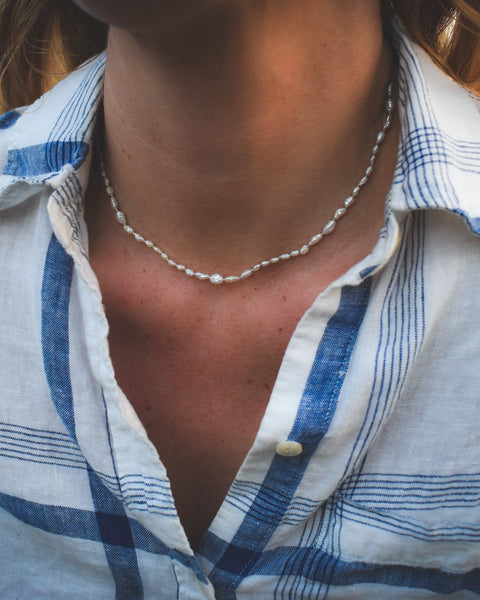 Person wearing a blue and white checkered shirt with a pearl necklace outdoors