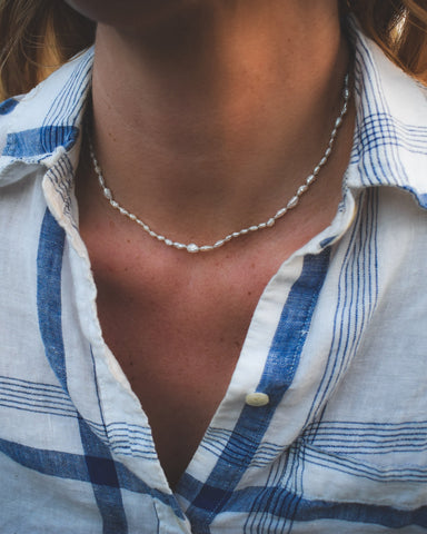 Person wearing a blue and white checkered shirt with a pearl necklace outdoors