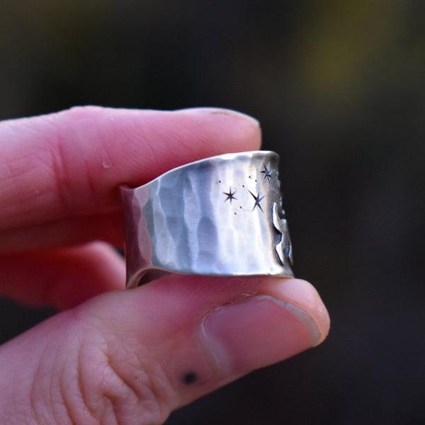 Silver ring with star and moon designs held between fingers against a blurred background