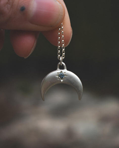 Silver crescent moon pendant with a blue gemstone held by a hand against a blurred background