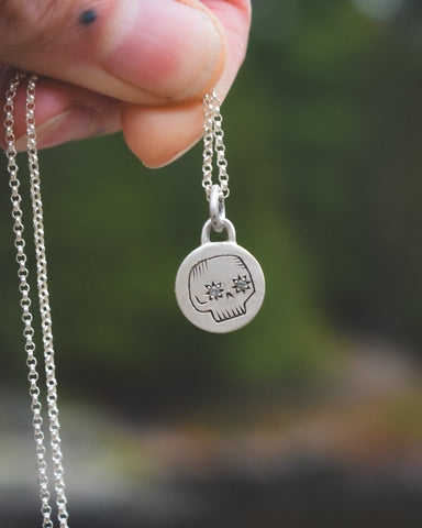 Silver necklace with a pendant featuring a face design held by a hand against a blurred natural background.